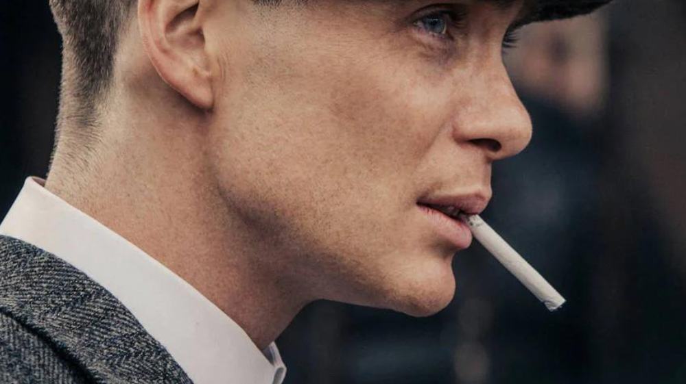 Cillian Murphy as Thomas Shelby in Peaky Blinders, wearing a flat cap and navy pinstripe suit before a train station. Quote: 'I’m not a traitor to my class... - Thomas Shelby.' Credit to @coscaexports.